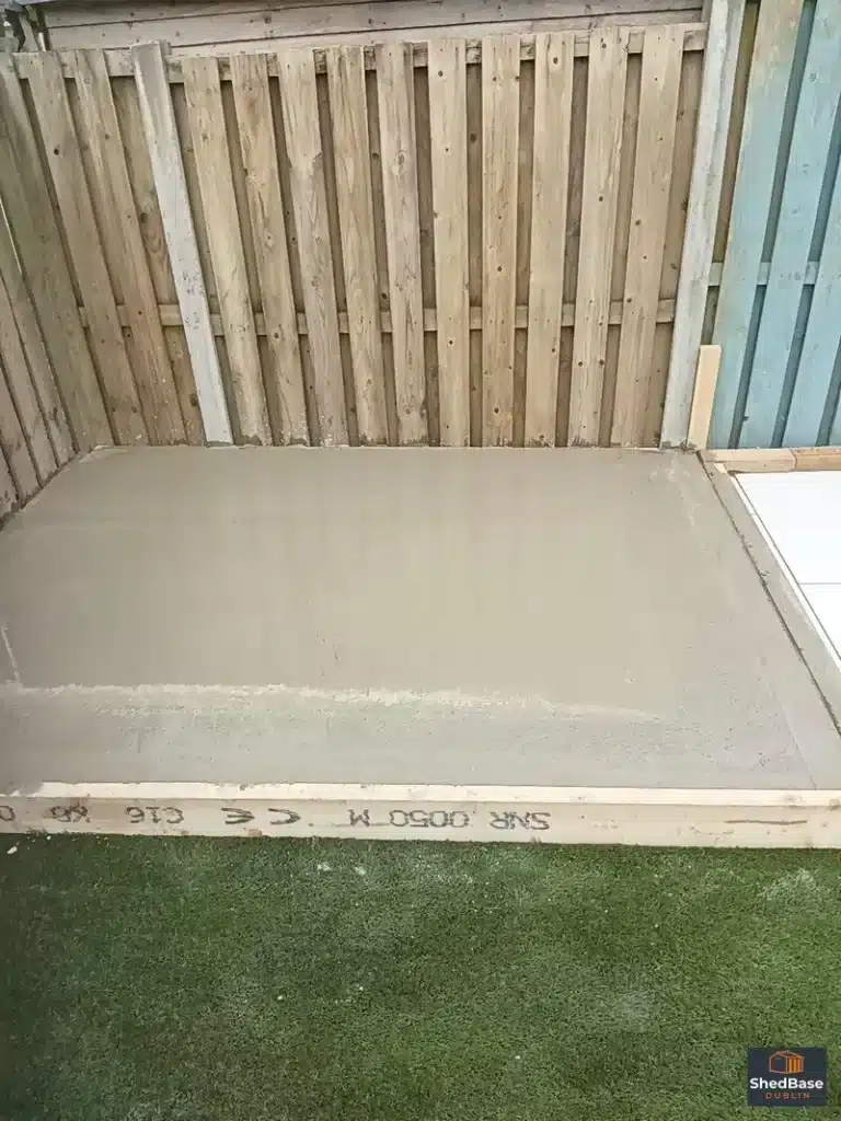 Fresh concrete shed base with timber formwork against a garden fence in Clontarf